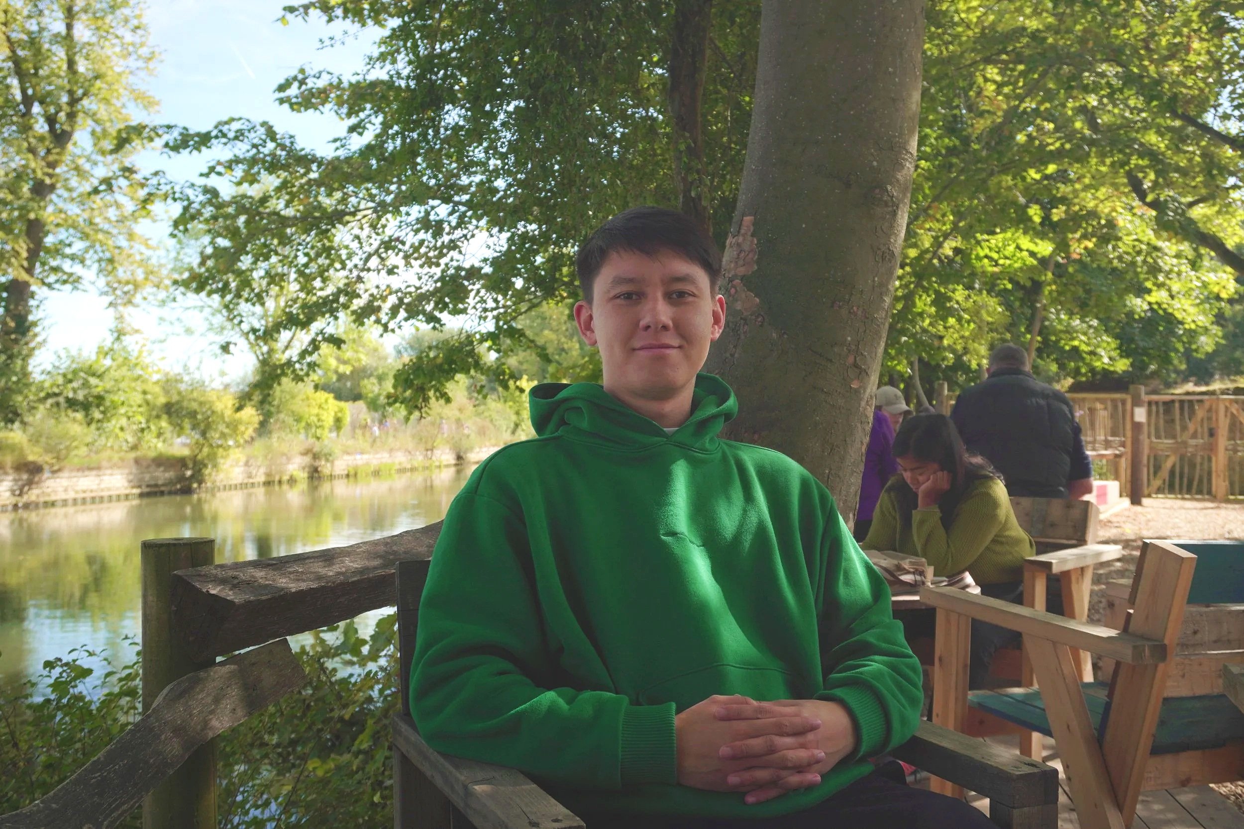 A young man in a green hoodie sitting outdoors by a riverside with trees, benches, and other people in the background.